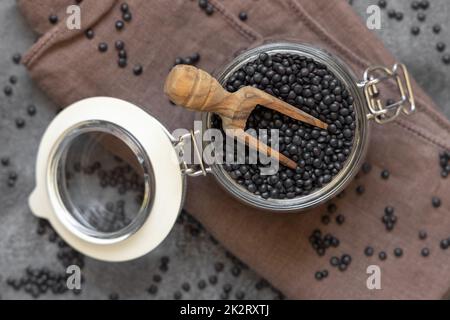 Glas Glas Glas Glas Glas aus trockenen schwarzen Linsen Bohnen mit einem Löffel auf grauem Tisch Blick, gesunde Protein Diät Stockfoto