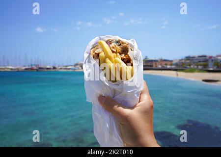 Pita Gyros. Griechische Gyros, eingewickelt in Pita-Brot, gegen den griechischen Strand mit blauem Meer. Stockfoto