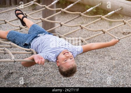 Fröhlicher, blonder Junge, der lächelt und auf einer Seilschaukel im öffentlichen Park liegt. Stockfoto