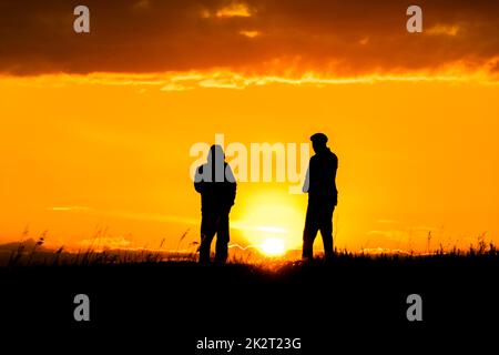 Zwei Silhouetten von Männern, die sich bei Sonnenuntergang oder Sonnenaufgang unterhalten, mit dramatischem Himmel und Wolken. Dialog und Begegnung mit zwei Menschen am Horizont und der Skyline.Umrisse von Menschen im Sonnenlicht.Goldene Stunde mit Freunden Stockfoto