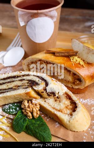 Mohn- und Apfelstrudel auf einem rustikalen Holztablett. Tee in einer Papiertasse mit einem weißen leeren Platz für Text und einem Löffel und einer Gabel aus umweltfreundlichen Materialien. Stockfoto