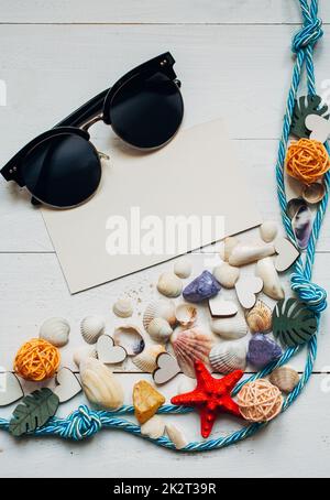 Muscheln, mehrfarbige Holzschmuck, Nautikschnur und Sonnenbrille auf weißem Holzhintergrund. Leere weiße Karte für Text. Stockfoto