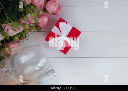Ein großer Strauß rosafarbener Rosen, ein Geschenk in einer roten Schachtel und eine Flasche Rosenwein mit Gläsern liegen auf der Seite auf einem weißen Holzhintergrund mit einem Platz für Text Stockfoto