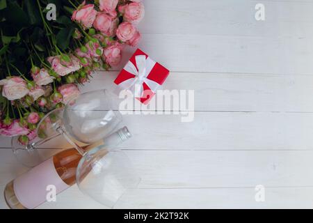 Ein großer Strauß rosafarbener Rosen, ein Geschenk in einer roten Schachtel und eine Flasche Rosenwein mit Gläsern liegen auf der Seite auf einem weißen Holzhintergrund mit einem Platz für Text Stockfoto