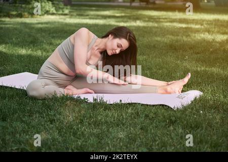 Eine junge Frau übt an einem warmen Sommertag Yoga in einem Stadtpark. Sportlerin sitzt auf einer Turnmatte. Stockfoto