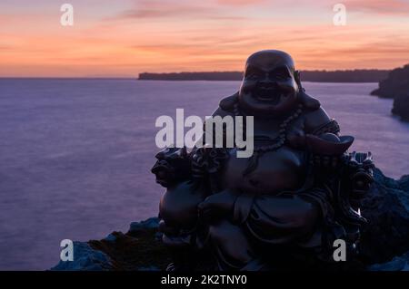 Buddha-Figur auf Felsen, bei Sonnenuntergang, auf dem Meer Stockfoto