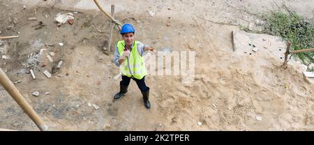 à¹à¹Young asiatischer Mann mit Schutzhelm und Sicherheitsweste, der auf ein Gerüst aus Holz zeigt, während er den Walkie-Talkie in der Hand hält. Sicherheitsüberprüfungen am Tag. Arbeitsumfeld am Standort von Wohnungsbauprojekten. Stockfoto