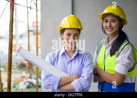 Junge Frau im Bauhelm und Grundriss mit gekreuzten Armen auf der Baustelle von Wohnbauprojekten. Asiatischer Ingenieur in einer Sicherheitsweste steht lächelnd neben ihr. Stockfoto