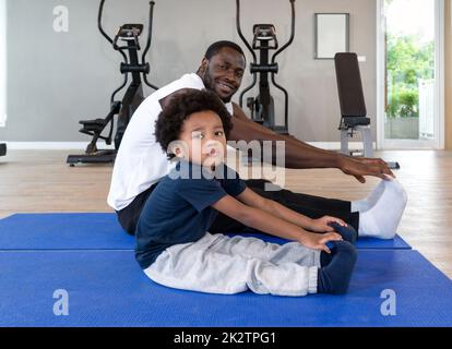 Junge lockige schwarze Haare Junge Stretching mit seinem Vater nach Abschluss der Übung Routine auf Yoga-Matte. Im Fitnessraum befinden sich im Hintergrund Ausdauergeräte. Stockfoto