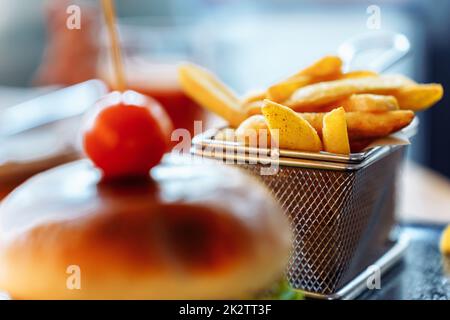 Konzentrieren Sie sich auf den Hintergrund von pommes frites in einem Gitterbehälter vor dem Hintergrund eines Burgers, der mit einer Kirschtomate dekoriert ist Stockfoto