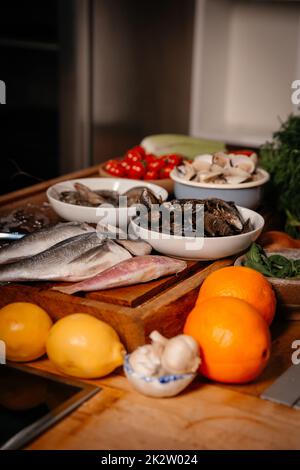 Auf dem Tisch neben Gemüse und Obst stehen Brassen, Knoblauch, Garnelen und Muscheln. Zutaten für die Zubereitung einer leckeren Suppe. Gute Küche. Stockfoto