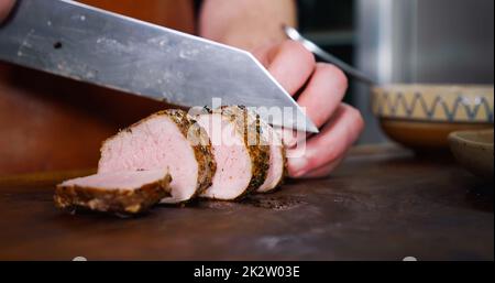 Der Koch schneidet saftiges Schweinesteak auf einem rustikalen Schneidebrett auf Holzhintergrund. Stockfoto