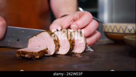 Der Koch schneidet saftiges Schweinesteak auf einem rustikalen Schneidebrett auf Holzhintergrund. Stockfoto