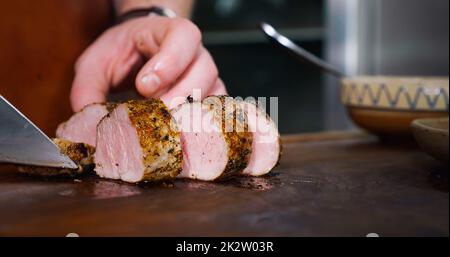 Der Koch schneidet saftiges Schweinesteak auf einem rustikalen Schneidebrett auf Holzhintergrund. Konzept der köstlichen Fleischgerichte. Nahaufnahme Stockfoto