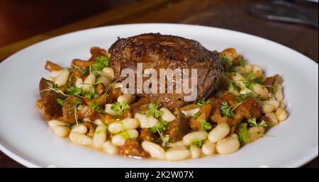 Professioneller Koch kocht in der Küche. Männliche Hände gießen köstliche Sauce auf Gericht mit Rindersteak und Bohnen. Stockfoto