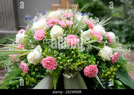 Pastellfarbene Trauerblumen auf einem Grab vor einem Holzkreuz in verschwommenem Hintergrund Stockfoto