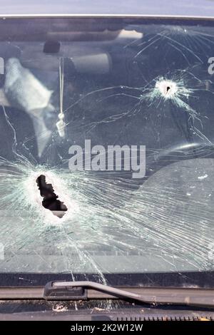 Löcher auf der Windschutzscheibe des Autos, es wurde von einer Waffe erschossen. Einschusslöcher. Zertrümmere die Windschutzscheibe, zerbrochenes und beschädigtes Auto. Die Kugel hat ein Loch im Glas zerbrochen. Stockfoto