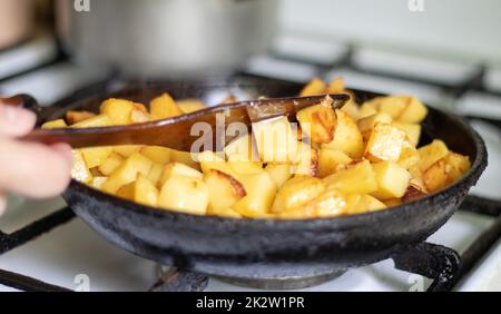 Braten frischer Kartoffeln in einer gusseisernen Pfanne mit Sonnenblumenöl. Blick auf eine Herdplatte mit einer Pfanne gefüllt mit goldenen Bratkartoffeln in einer echten Küche. In einer hausgemachten Bratpfanne gekochtes Essen. Stockfoto