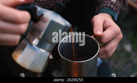 Ein junges Paar im Wald. Frau gießt Tee, Kaffee aus einer Kaffeemaschine aus Aluminium ein. Ein Campingpaar, das Kaffee in eine Tasse gießt. Stockfoto