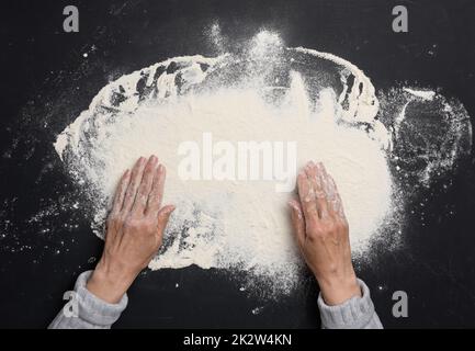 Gesiebt Weizenmehl auf einem schwarzen Tisch und zwei weibliche Hände, Draufsicht. Kochen zu Hause Stockfoto