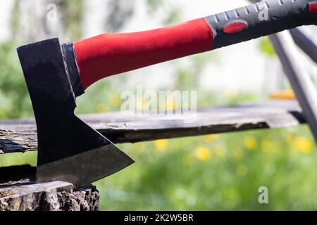 Eine neue moderne Axt mit rotem Griff ragt an einem sonnigen Sommertag aus einem Holzstumpf auf einer grünen Wiese hervor. Die Axt steckte im Stumpf fest. AX-Klinge in einem Baumstamm. Stockfoto