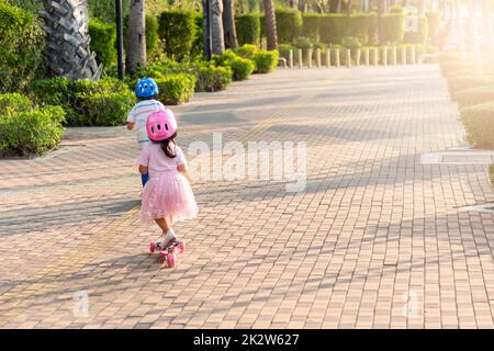 Zurück asiatische kleine Jungen Jungen und Mädchen tragen Sicherheitshelm spielen Kickboard auf der Straße im Park draußen am Sommertag Stockfoto