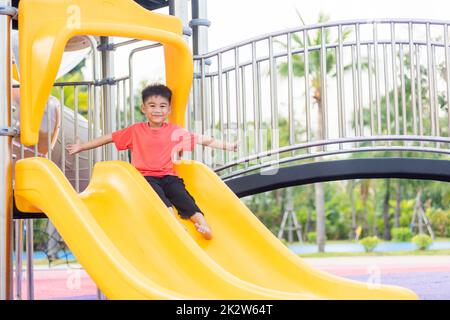 Ein asiatisches Kind lächelt auf dem Spielplatz mit Schiebebügel, ein glückliches Kind im Vorschulalter, das tagsüber im Sommer auf der Spielplatzausrüstung spielt, Klettern eines kleinen Jungen Stockfoto