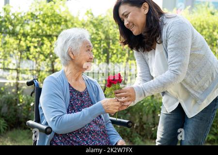 Altenpfleger Tochter umarmen und helfen asiatische ältere oder ältere alte Dame Frau mit roten Rose auf Rollstuhl im Park. Stockfoto