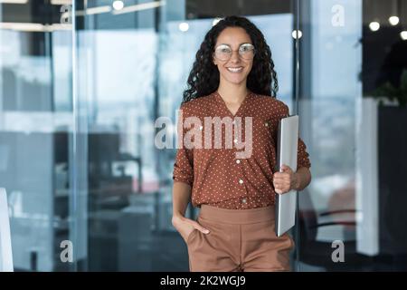 Porträt einer jungen schönen hispanischen Studentin mit Laptop, einer Frau bei der Arbeit im modernen Universitätsklassenzimmer, lächelnd und mit Blick auf die Kamera, einer Lehrerin in Brille und lockigen Haaren. Stockfoto
