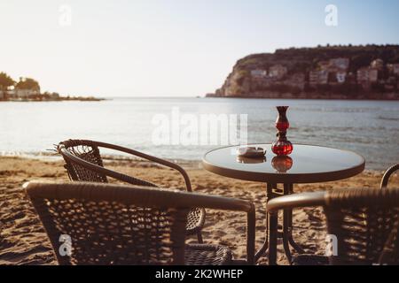 Rückansicht Nahaufnahme von leerem Kaffeetisch und Stühlen am Sandstrand mit Aschenbecher und Kerzenlicht vor dem Golf am Meer mit klarem Himmel vor Sonnenuntergang Stockfoto
