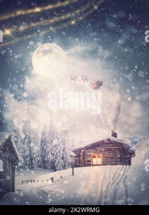 Zauberhafte Urlaubsszene und Weihnachtsmannschlitten mit Rentieren, die am Heiligabend über dem verschneiten Haus fliegen. Wunderbare Schneeflocken über der Villa Stockfoto