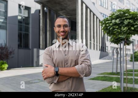 Junger erfolgreicher Geschäftsmann lächelt und schaut mit gekreuzten Armen auf die Kamera, Mann vor dem Bürogebäude trägt Hemd und Brille, Startup-Investor. Stockfoto