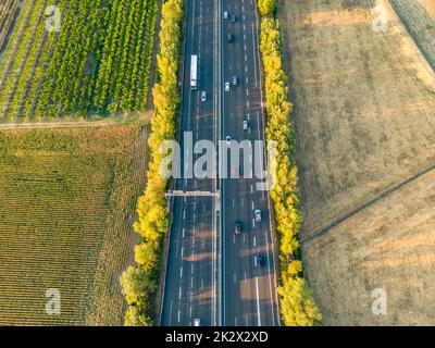 Autobahn aus der Vogelperspektive. Wenn die Sonne untergeht, erzeugen die Sonnenstrahlen wunderschöne lange Schatten auf dem Boden. Gelbe Bäume und umliegende Farmlan Stockfoto