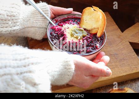 Weibliche Hände halten eine Schüssel Gemüsesuppe mit Rote Beete, Käse, Sauerrahm und Brot auf arustischem Holzhintergrund. Stockfoto