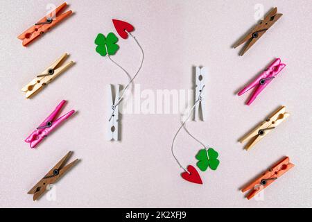 Farbenfrohes Set aus verschiedenen Holz- und Kunststoffklammern auf einem hellrosa glitzernden Hintergrund. Ein rotes und ein grünes Glückkleeblatt sind an jedem der weißen Holzklammern befestigt. Draufsicht. Stockfoto