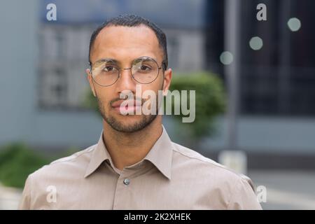 Nahaufnahme eines Portraits eines jungen Geschäftsmannes im Hemd, eines ernsthaften und nachdenklichen Mannes, der vor dem modernen Bürogebäude eine Brille trägt und die Kamera ansieht. Stockfoto
