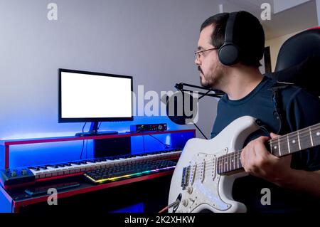 Der junge lateinische Kaukasusmann mit Bart und Brille sitzt in seinem Heimmusikstudio und überprüft die Aufnahme, die er mit seiner E-Gitarre gemacht hat, beim Einchecken Stockfoto