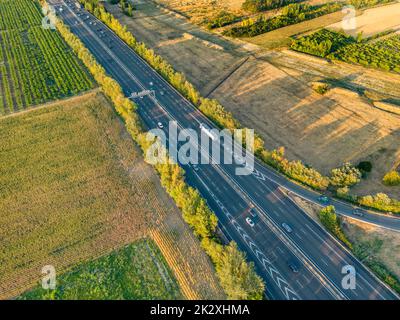 Autobahn aus der Vogelperspektive. Wenn die Sonne untergeht, erzeugen die Sonnenstrahlen wunderschöne lange Schatten auf dem Boden. Gelbe Bäume und umliegende Farmlan Stockfoto