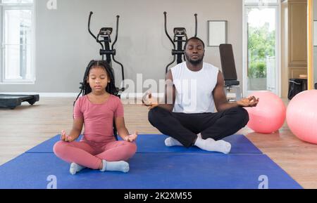 à¸Black Cornrow Braids Mädchen in Sportbekleidung meditiert mit ihrem Vater auf Yogamatte vor dem Training. Zwei Yoga-Bälle werden daneben platziert. Morgengymnastik, Achtsamkeitskonzept. Stockfoto