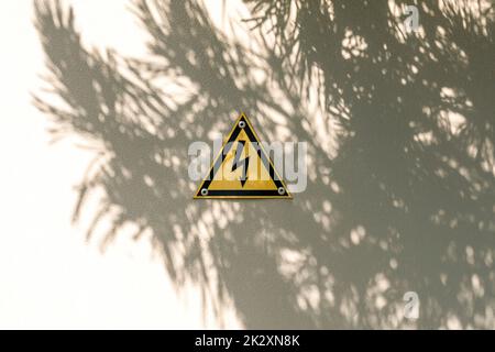 Warnschild mit gelbem Hochspannungsdreieck an grauen Metalltüren mit Baumschatten Stockfoto