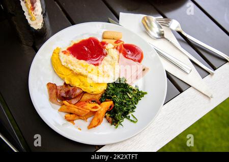 Frühstück vom Buffet auf einem runden weißen Teller. Auf einem modernen Holztisch steht ein Glas Wasser auf dem Kopf, das reflektiert. Daneben befinden sich Edelstahlmesser, Löffel und Gabeln. Stockfoto