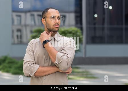 Junger, nachdenklicher Geschäftsmann mit Brille vor dem modernen Bürogebäude, Mann, der denkt und wegschaut, Büroangestellter im Hemd. Stockfoto