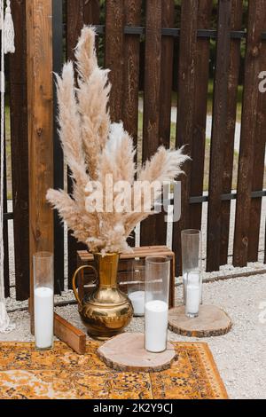 Hohe Pampa Gras in einer Vase und Kerzen in Glasbirnen gegen einen Holzzaun in Dekoration Party. Pampas Gras im Innenraum, Boho-Stil. Dekor für Stockfoto