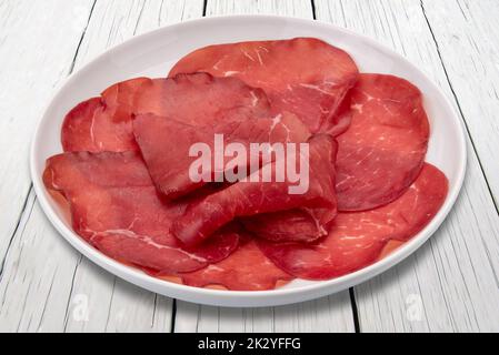 Brosaola Scheiben in Teller auf weißem Holztisch, italienische getrocknete Rindersalami aus Valtellina Stockfoto