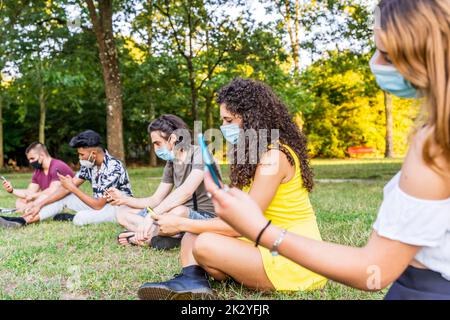 Eine Gruppe von multirassischen Freunden, die in einem öffentlichen Park im Freien sitzen und mit Smartphones Inhalte in sozialen Netzwerken teilen Stockfoto
