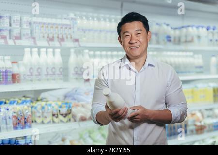 Porträt eines glücklichen und zufriedenen asiatischen Käufers im Supermarkt in der Milchabteilung, eines Mannes, der lächelnd eine Flasche Milch in der Hand hält und in der Nähe von Kühlschränken auf die Kamera schaut Stockfoto