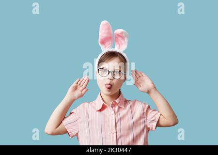 Portrait eines Teenagers mit Down-Syndrom, das Osterhasen-Ohren vor blauem Hintergrund trägt und lustige Gesichter macht Stockfoto