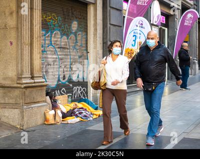 Ein Ehepaar mit Gesichtsmasken auf der Straße, das an einem Obdachlosen vorbeigeht, der im Zentrum von Sol in Madrid, Spanien, schläft Stockfoto