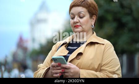 Eine elegante Frau mit kurzen roten Haaren und einem Regenmantel steht auf der Straße und benutzt ein Smartphone Stockfoto