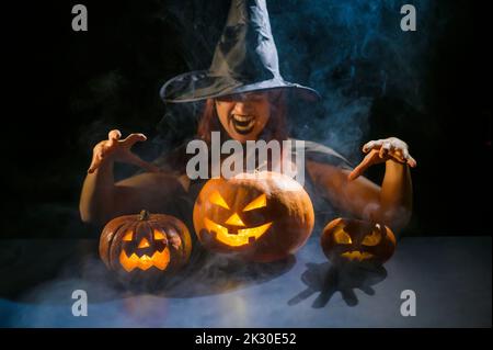 Eine ominöse Hexe im Hut zaubert über eine Jack-o-Laterne. Traditionelle halloween-Figuren. Mystischer Nebel kriecht über Kürbisse mit geschnitzten schrecklich Stockfoto